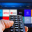 A man is holding a remote control of a smart TV in his hand. In the background you can see the television screen with streaming entertainment apps for video on demand