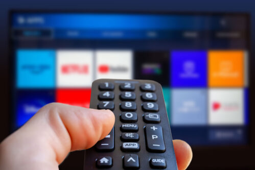 A man is holding a remote control of a smart TV in his hand. In the background you can see the television screen with streaming entertainment apps for video on demand A man is holding a remote control of a smart TV in his hand. In the background you can see the television screen with streaming entertainment apps for video on demand