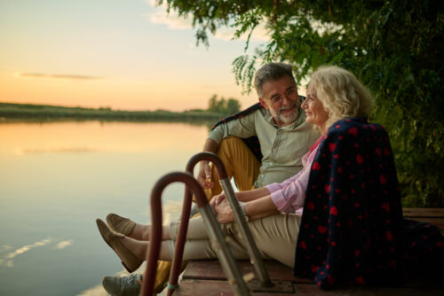 Elderly couple is enjoying a peaceful evening on a wooden pier, sharing a blanket and watching the sunset over the lake